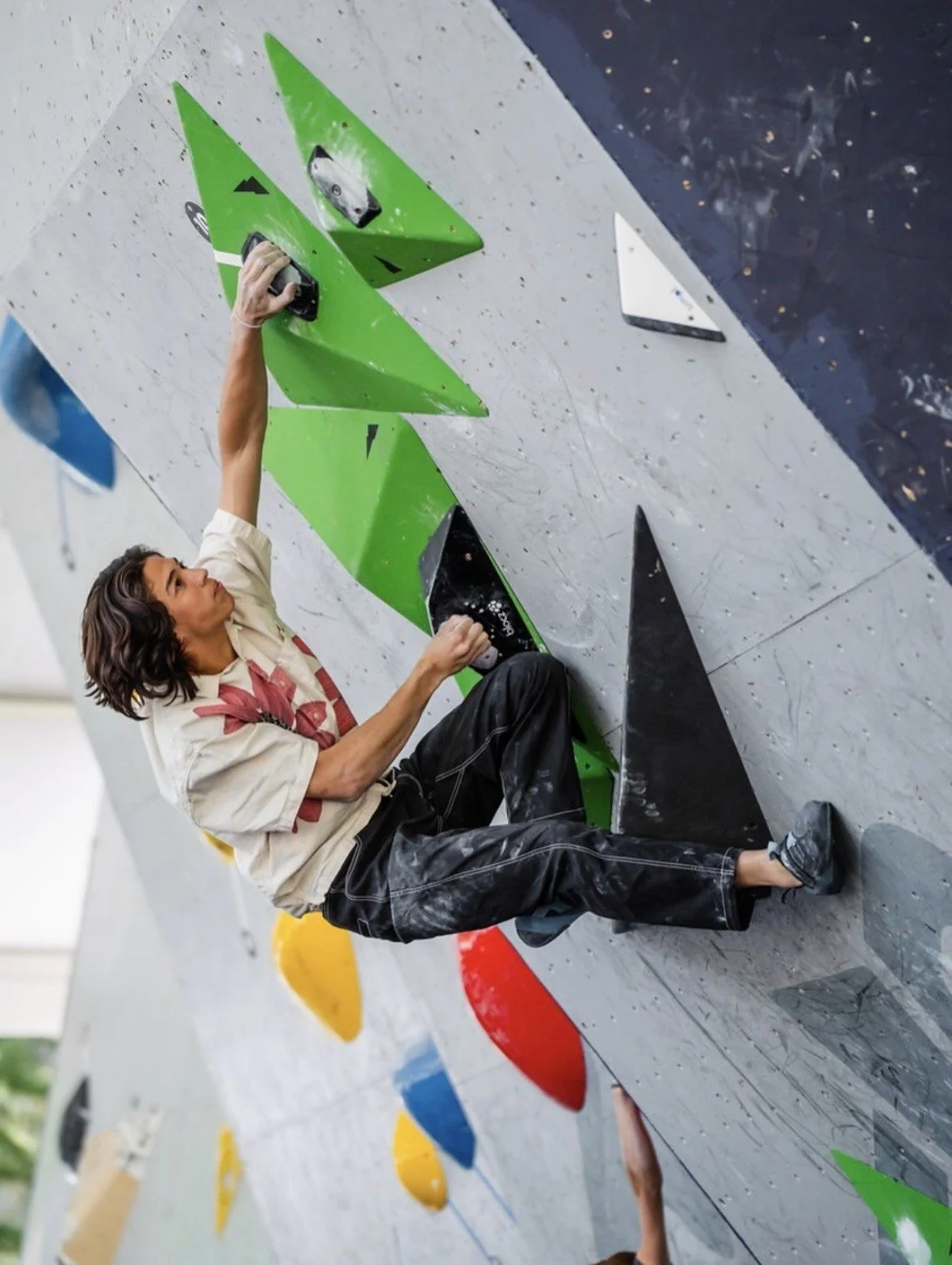 Young person with long hair climbing an indoor rock wall, holding onto green holds, wearing a beige shirt with a red design and black pants.