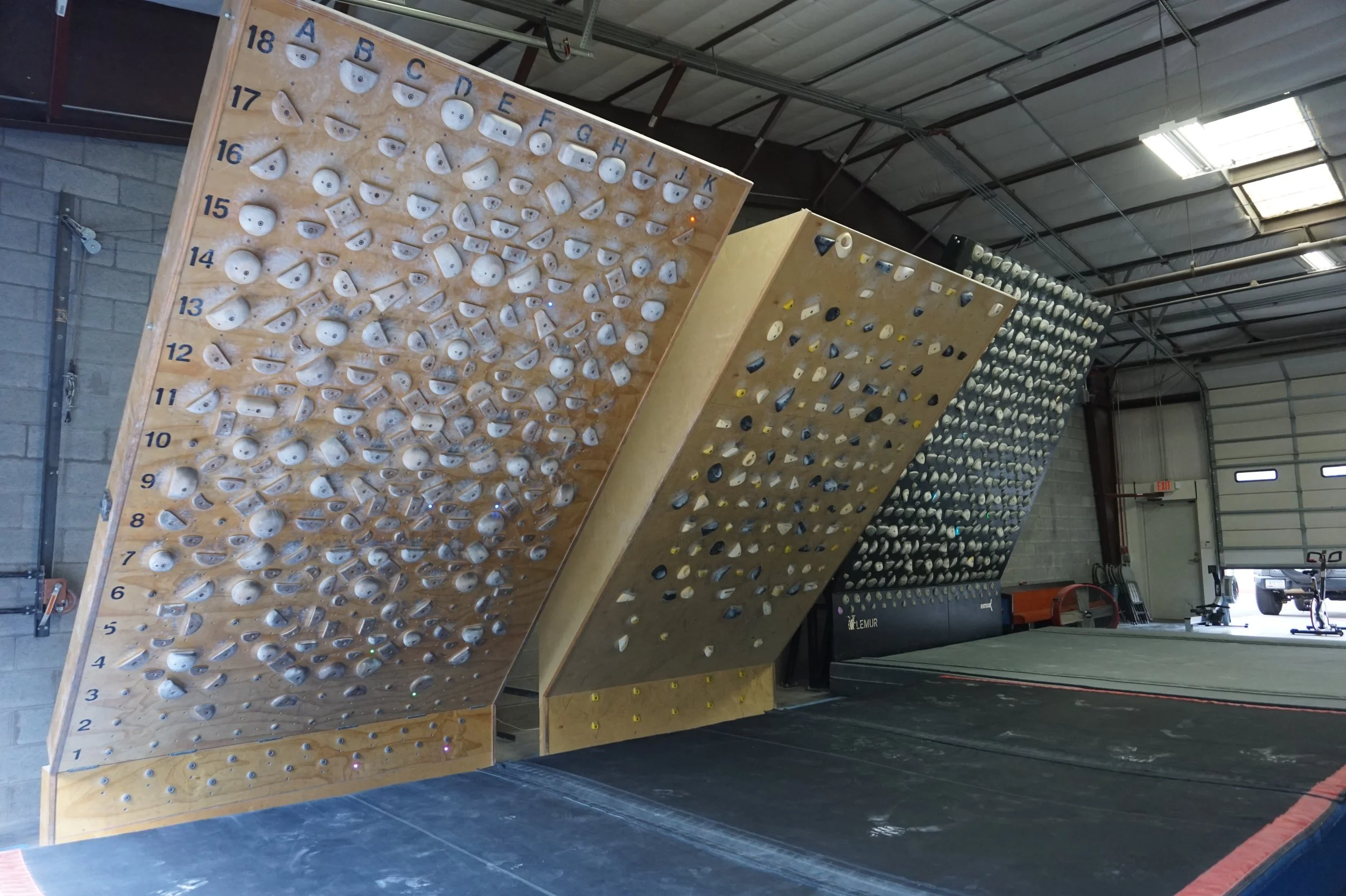 Indoor rock climbing gym with several climbing walls equipped with numerous holds and ledges for climbers to grip and step on, within a spacious industrial-style building.