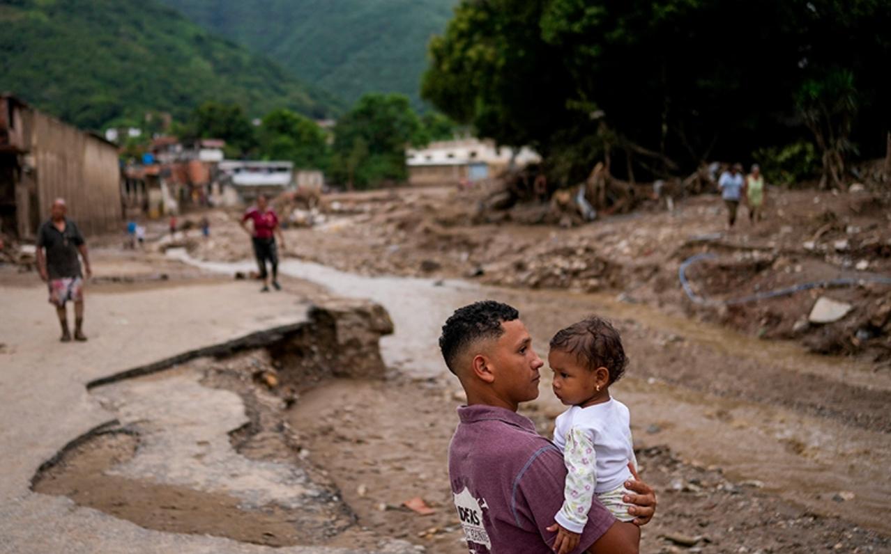 A man holding a young girl in a damaged and muddy area with collapsed infrastructure and other people walking in the background, surrounded by green hills and trees.
