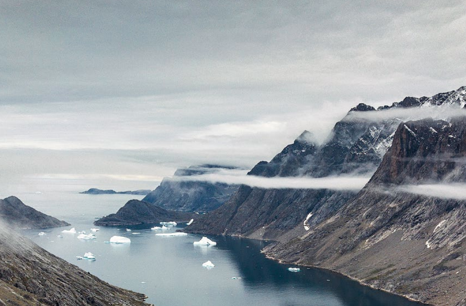 A fjord with steep mountains on either side, snow-capped peaks, and small icebergs floating in the water.