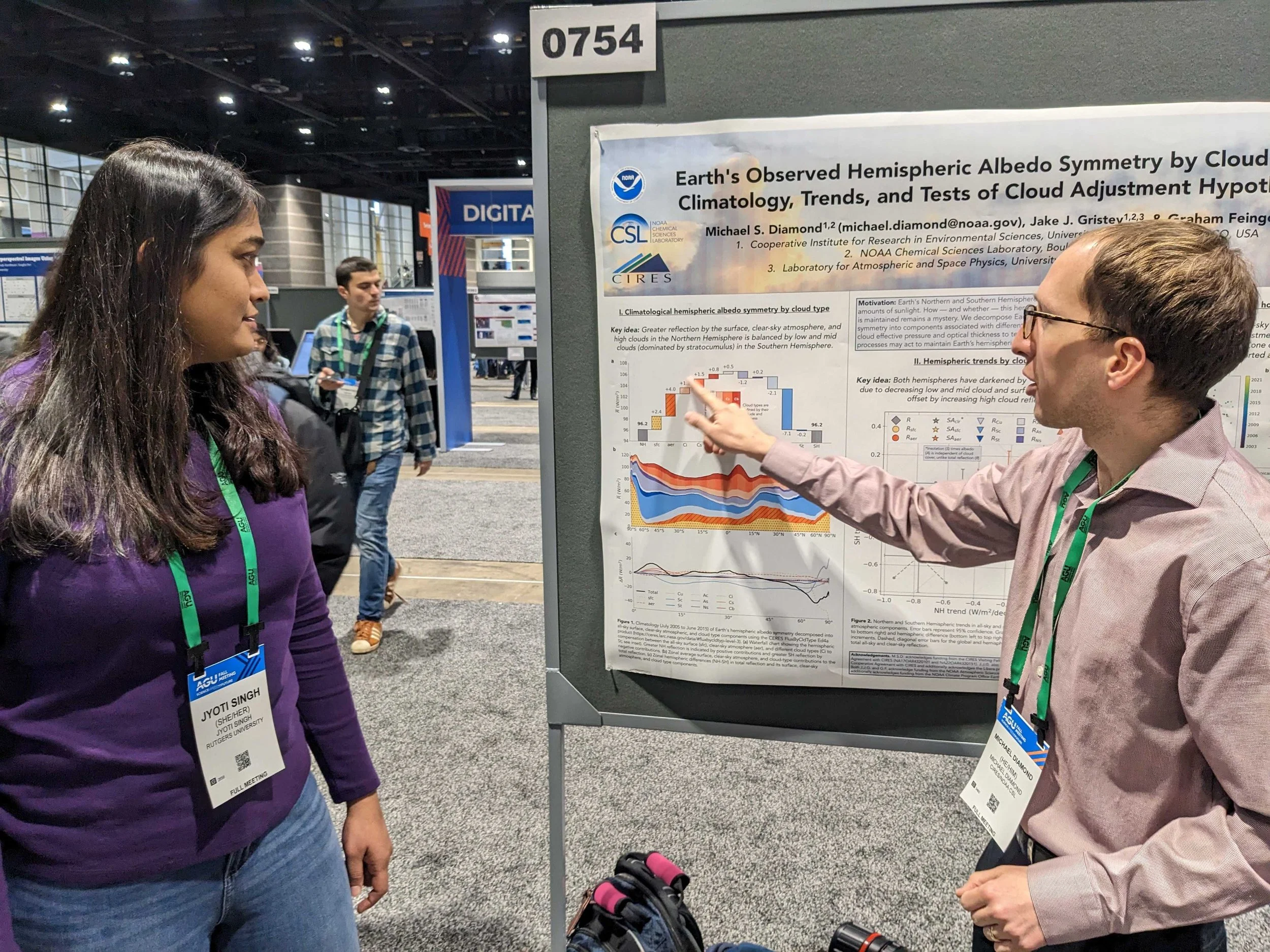 A man is presenting a scientific research poster at a conference while a woman listens. The poster discusses Earth's hemispheric albedo symmetry and climate trends. The man is pointing at the poster, and both are wearing conference badges.