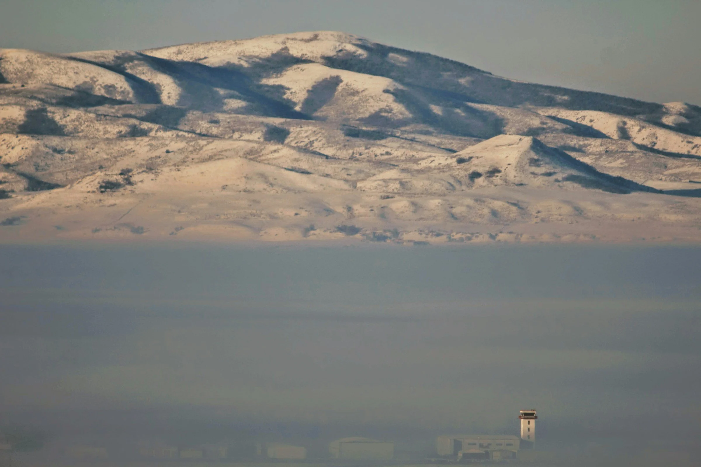 Snow-covered mountains in the background with a layer of fog at the base, and an airport control tower and buildings in the foreground.