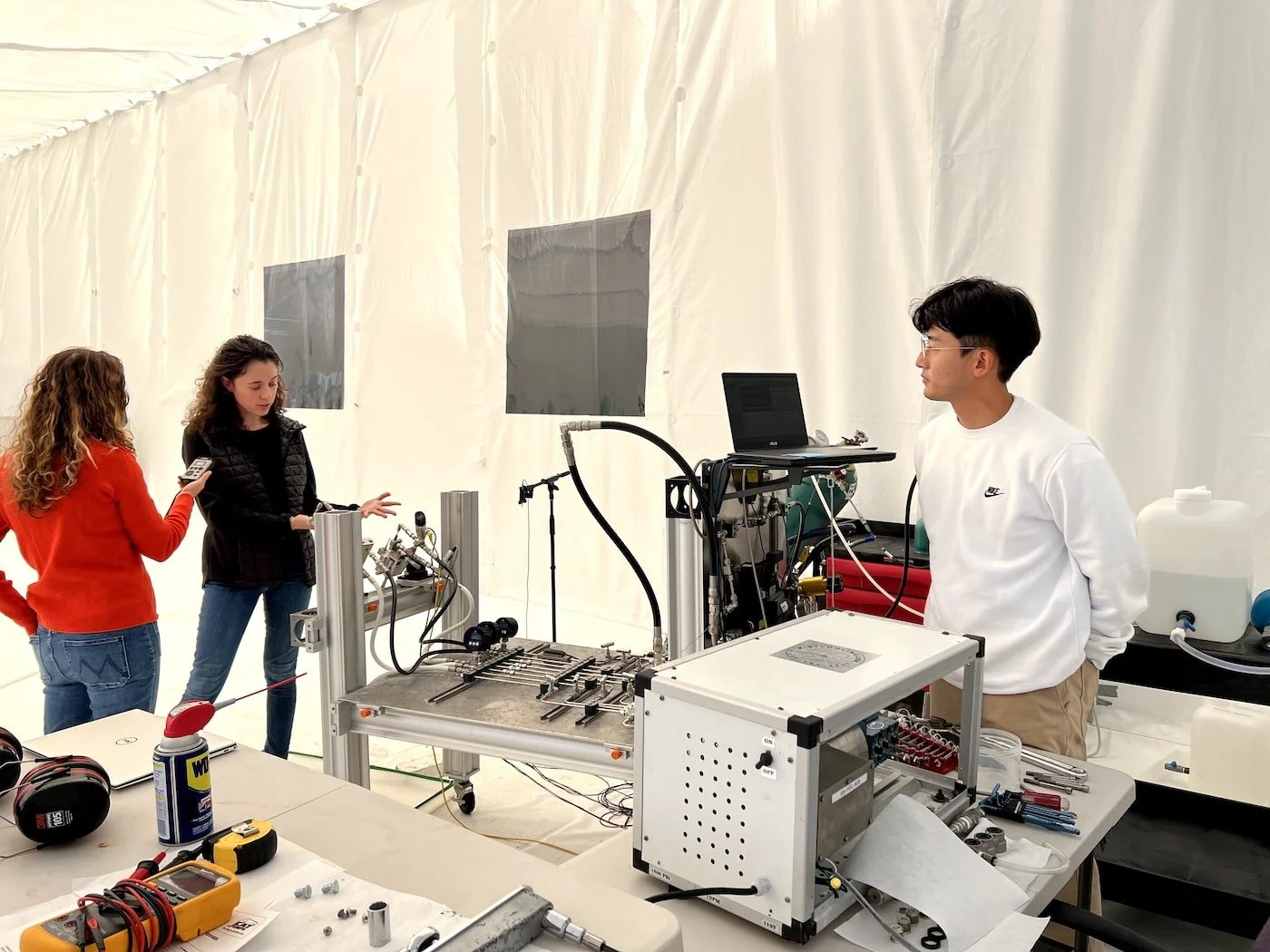 Three women and one man in a laboratory with technical equipment; the women are discussing, and one woman is holding a smartphone.