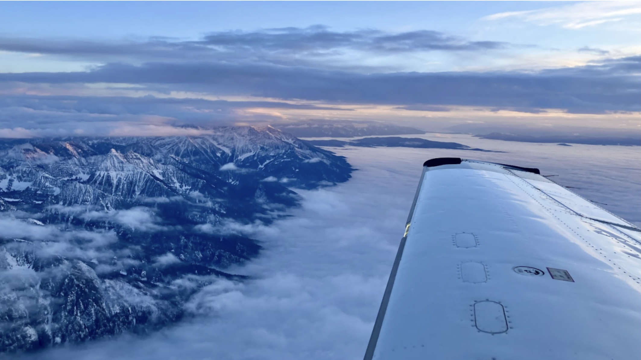 View from an airplane window showing snow-capped mountains, clouds, and a river or lake below, with the airplane wing visible on the right.
