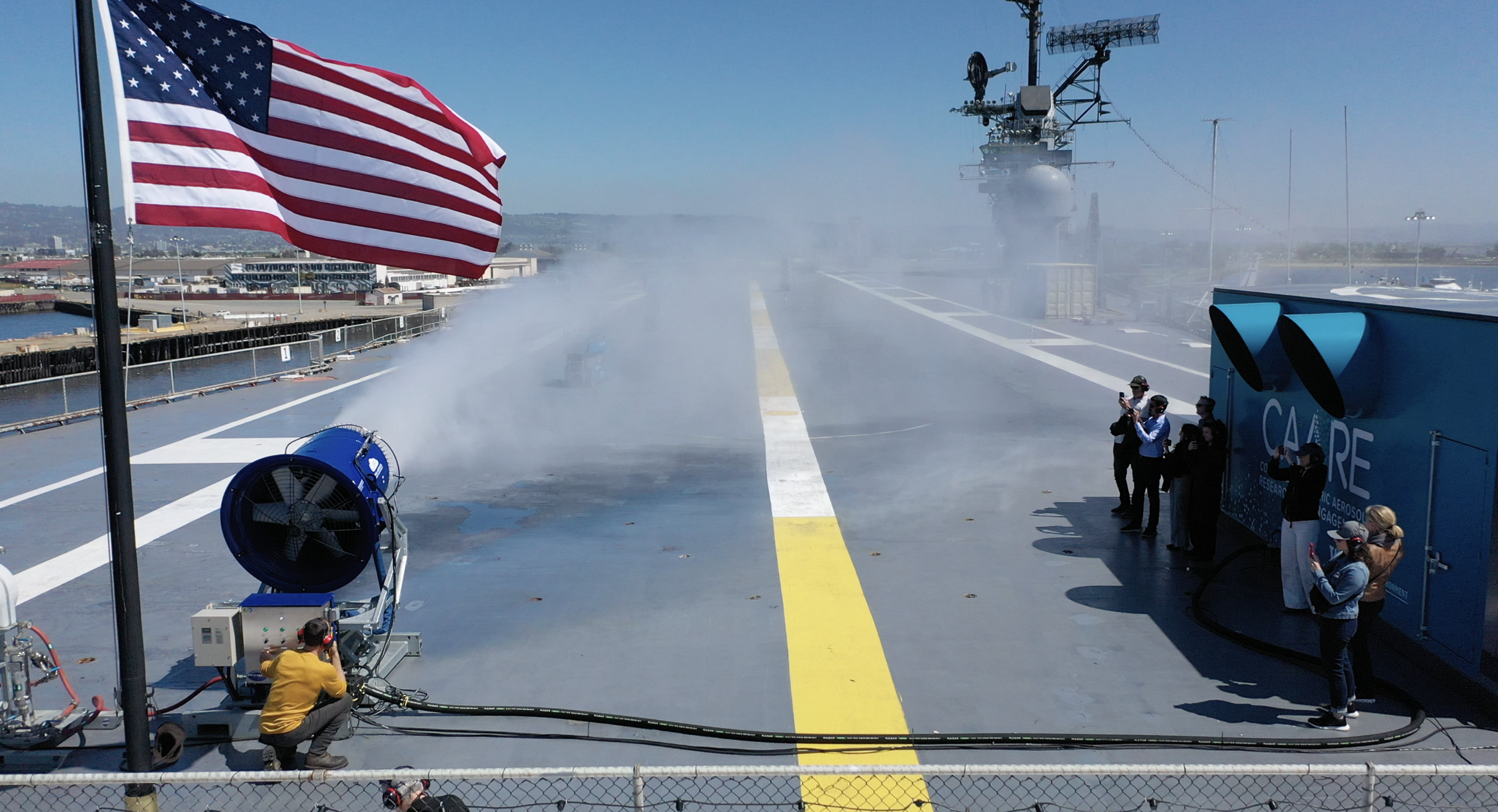 On the deck of an aircraft carrier, a large American flag flies, and a crew member operates a fog dispersal machine, creating a cloud of mist. Several onlookers stand nearby, some taking photos, as the ship's radar and communication equipment are visible in the background.