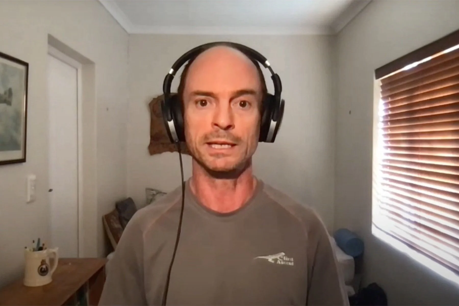 A man with a receding hairline wearing large over-ear headphones and a gray T-shirt stands in a room with white walls, wooden blinds, and a small wooden table with a mug on it.