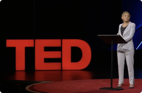 A woman in a white suit standing at a podium on a stage with a large red TED logo behind her.