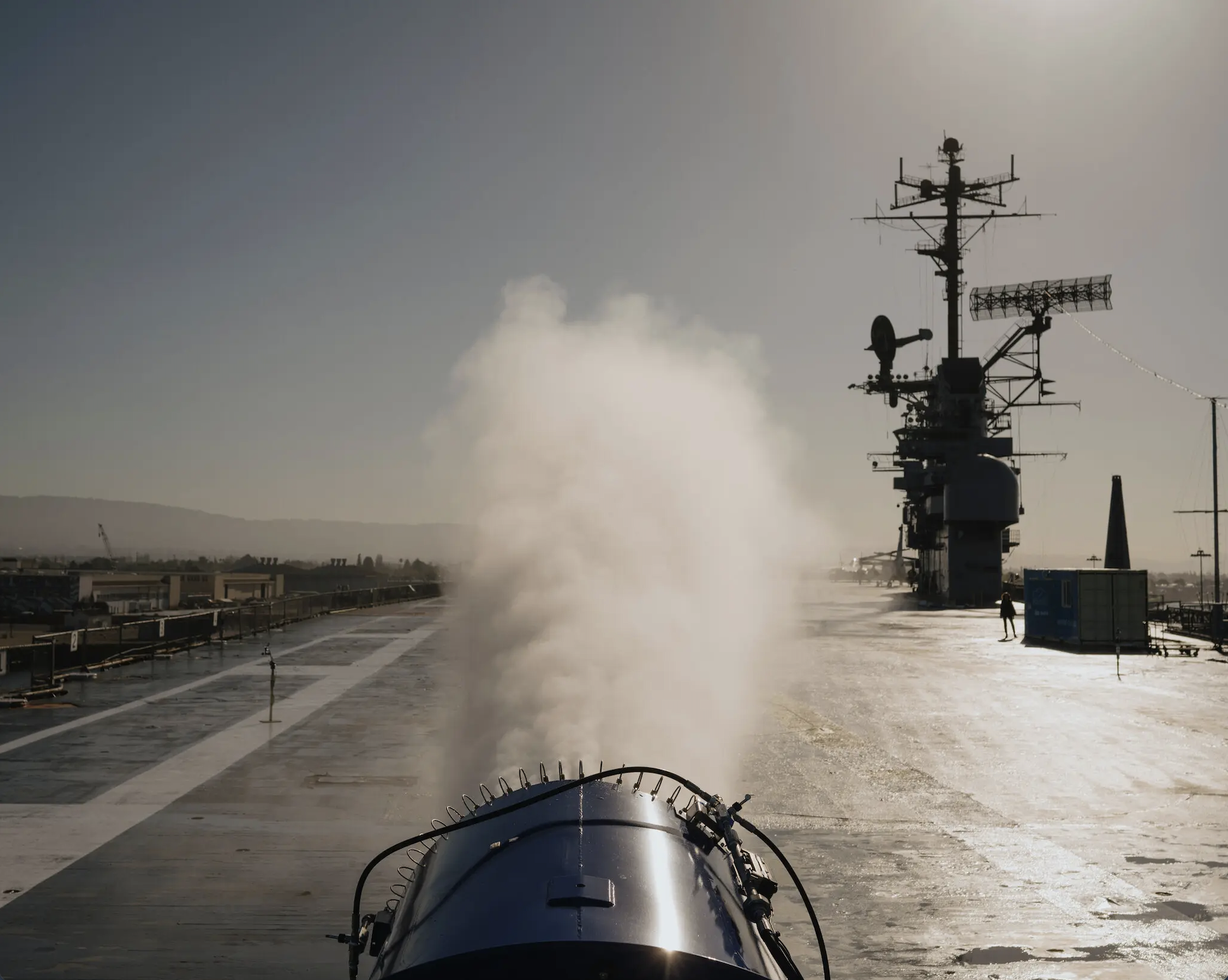 View from the deck of an aircraft carrier showing a large gun firing, with smoke rising, and the ship's radar and communication equipment in the background.