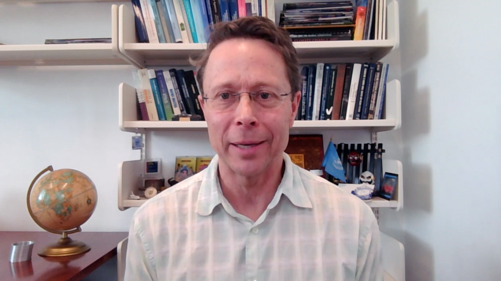 A man wearing glasses and a white plaid shirt sitting in front of a desk with a globe on it, in an office with bookshelves filled with books and small decorations behind him.