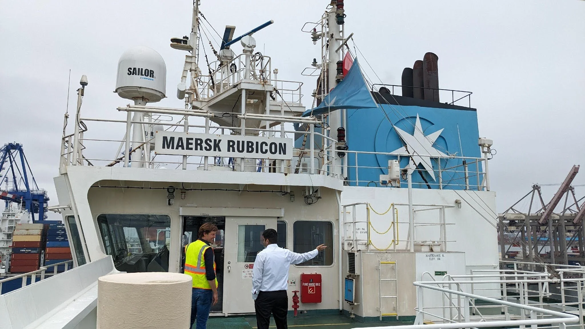 A white cargo ship named Maersk Rubicon docked at a port with cranes in the background. Two men are standing near the ship's entrance, one explaining something to the other. The ship's logo and various equipment are visible on the superstructure.