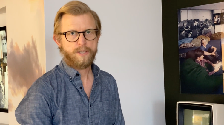 A man with glasses and a beard wearing a denim shirt, standing indoors near a black wall with photographs and a vintage television.