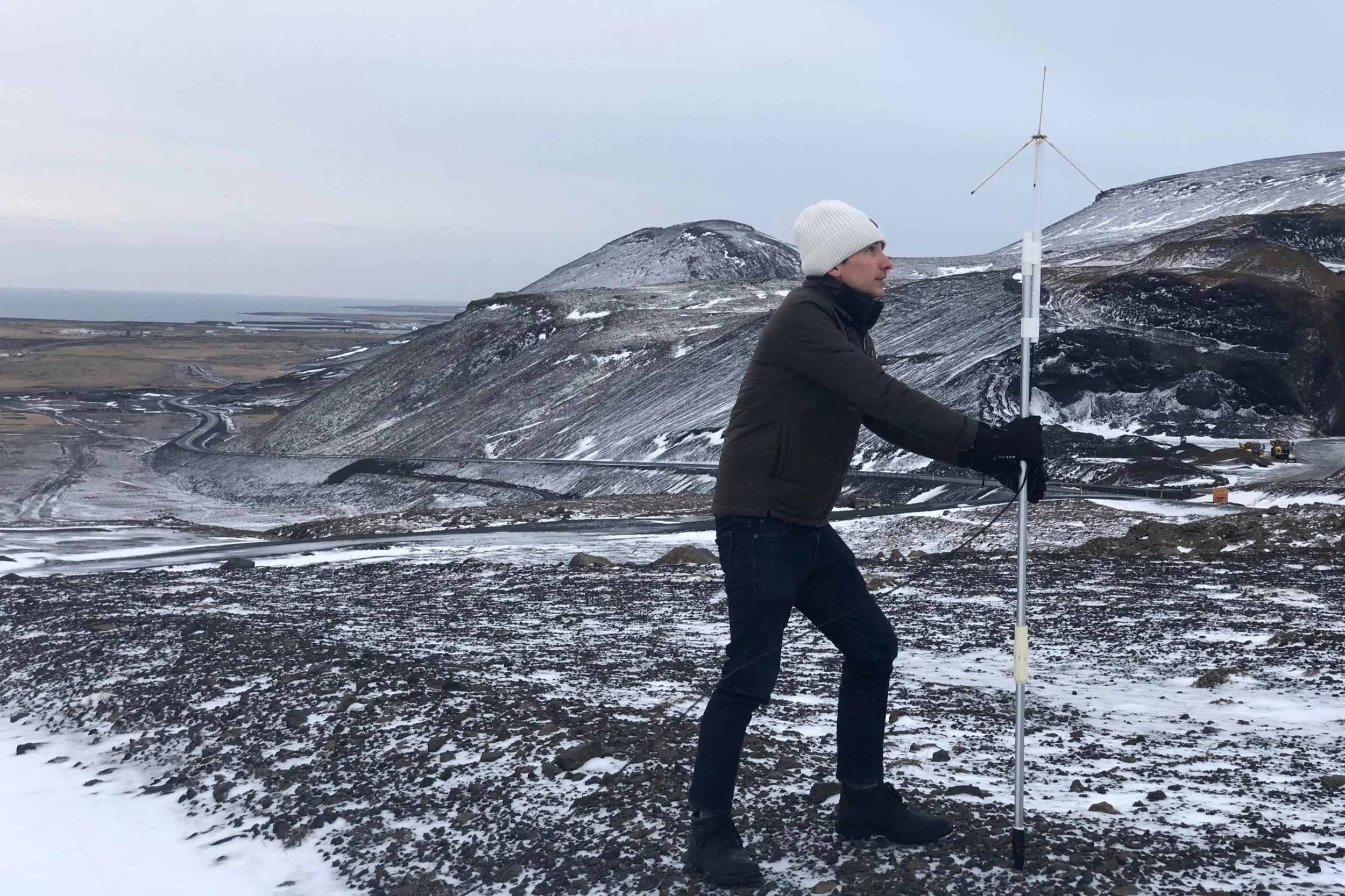 A person in winter clothing stands outdoors on rocky terrain with snow, holding a pole with a wind turbine model, in a cold, mountainous landscape.