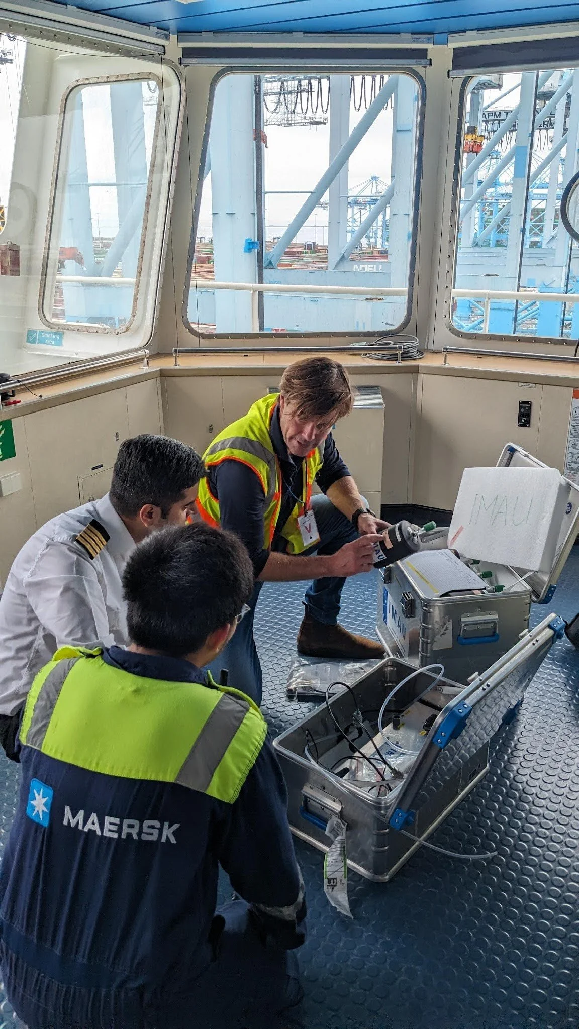 Four men, two in high visibility jackets and one in a pilot uniform, are inspecting equipment inside an aircraft cockpit. One man is holding a flashlight, and the others are observing. The setting appears to be an inspection or maintenance operation at an airport or ferry terminal.