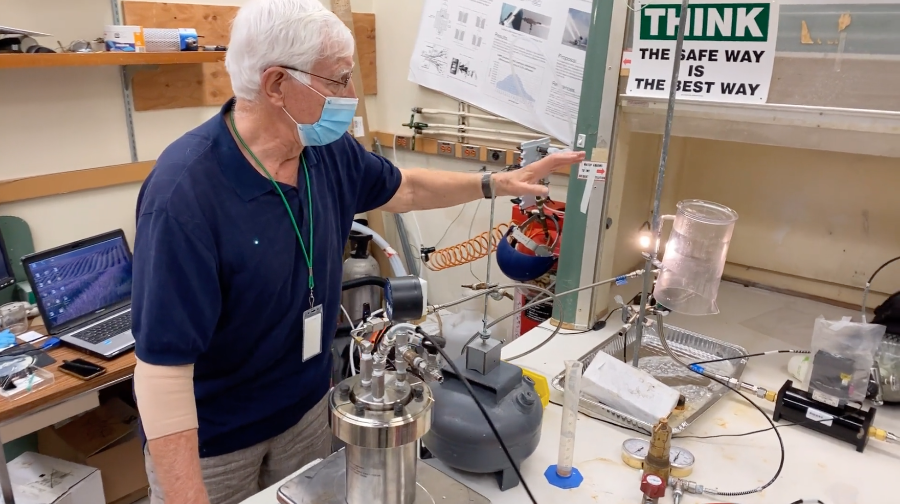 A man wearing a face mask and glasses is conducting a scientific experiment in a laboratory with various glassware, tubes, and equipment. There is a laptop on the desk behind him and safety posters on the wall.