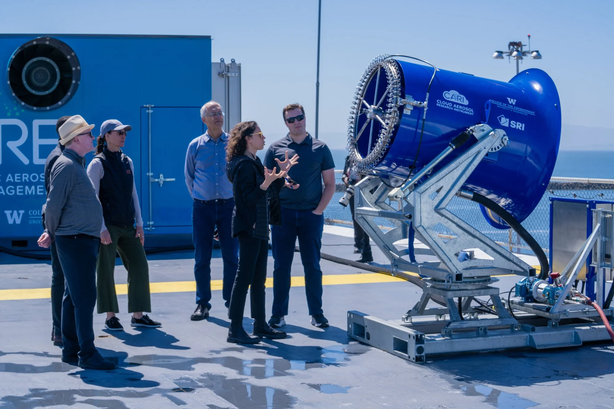 A group of people on a rooftop observing a large scientific instrument, which is a blue cylindrical device mounted on a metal stand, part of an aerospace research setup with a shipping container in the background.