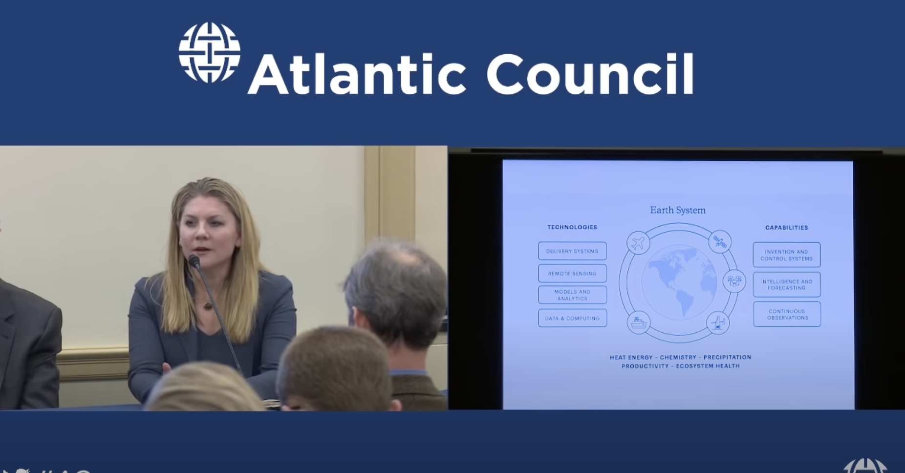 A woman with blonde hair, speaking into a microphone during a panel at an Atlantic Council event. Behind her, a large screen displays a presentation slide titled 'Earth System' with diagrams and list of technologies and capabilities related to Earth systems.