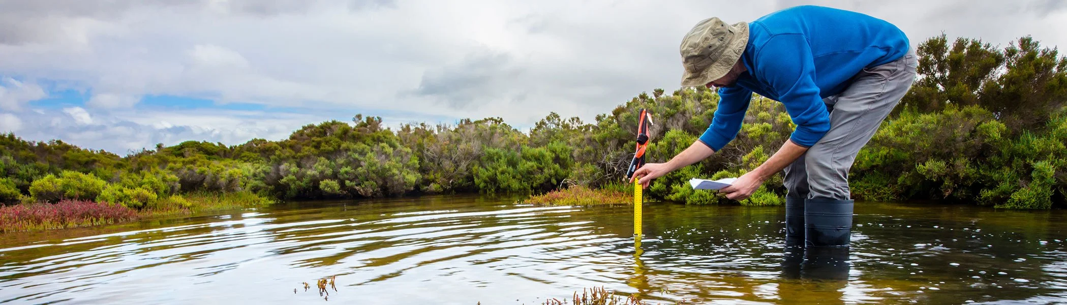A person wearing a beige hat, blue shirt, gray pants, and black boots leaning over a riverbank, taking measurements with a yellow measuring tape and holding a clipboard under a cloudy sky.