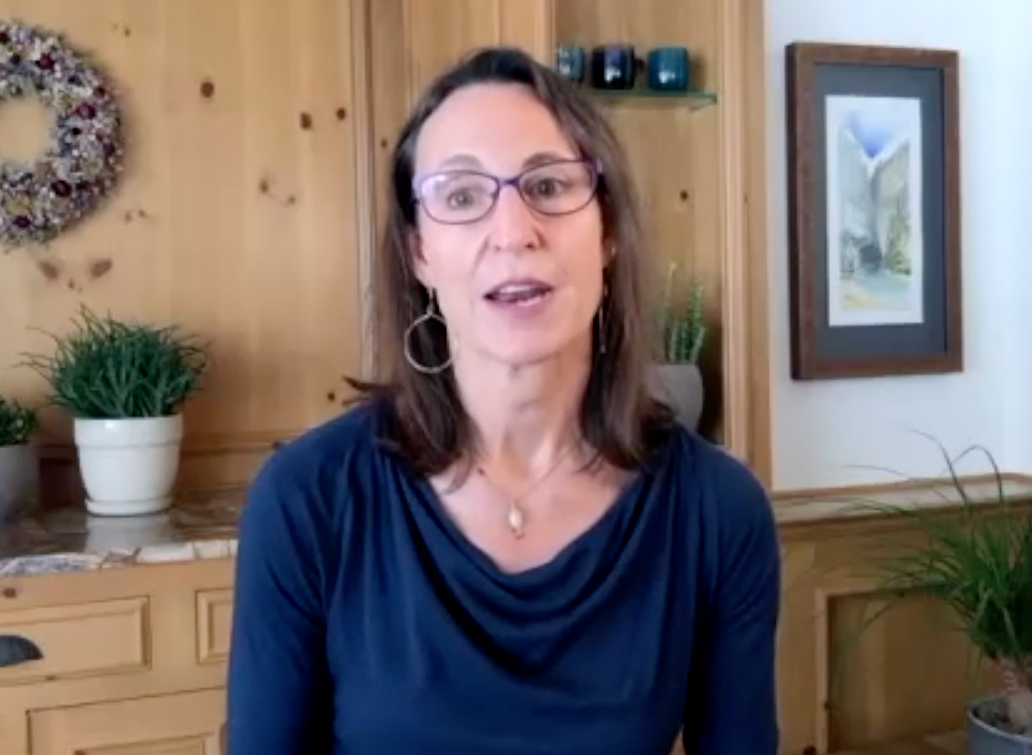 Woman with shoulder-length brown hair, glasses, wearing a dark blue top, sitting or standing in a room with wooden walls and plants, speaking to the camera.