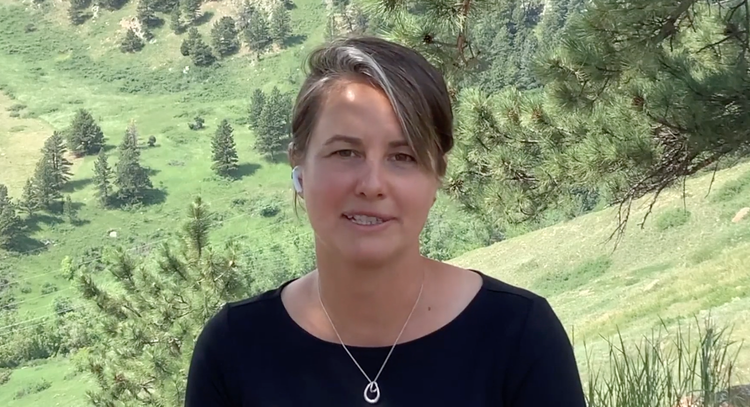 A woman outdoors in a green, hilly, forested area with trees and grass, wearing a black shirt, necklace, and earbuds.