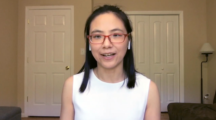 Woman wearing glasses and wireless earbuds sitting in a room with two closed doors, a gray couch, and a floor lamp.