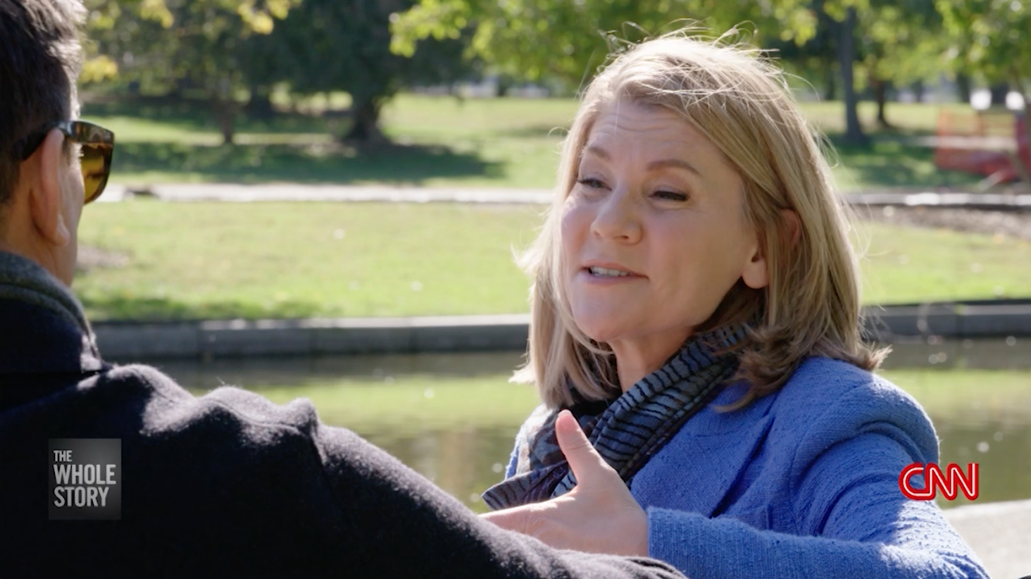 A woman and a man having a conversation outdoors in a park. The woman is facing the camera, smiling, and wearing a blue jacket with a patterned scarf. The man, seen from behind, is wearing sunglasses and a dark jacket. They are near a pond with trees in the background. The CNN logo is in the bottom right corner, and a black box with white text reads "The Whole Story" in the bottom left corner.