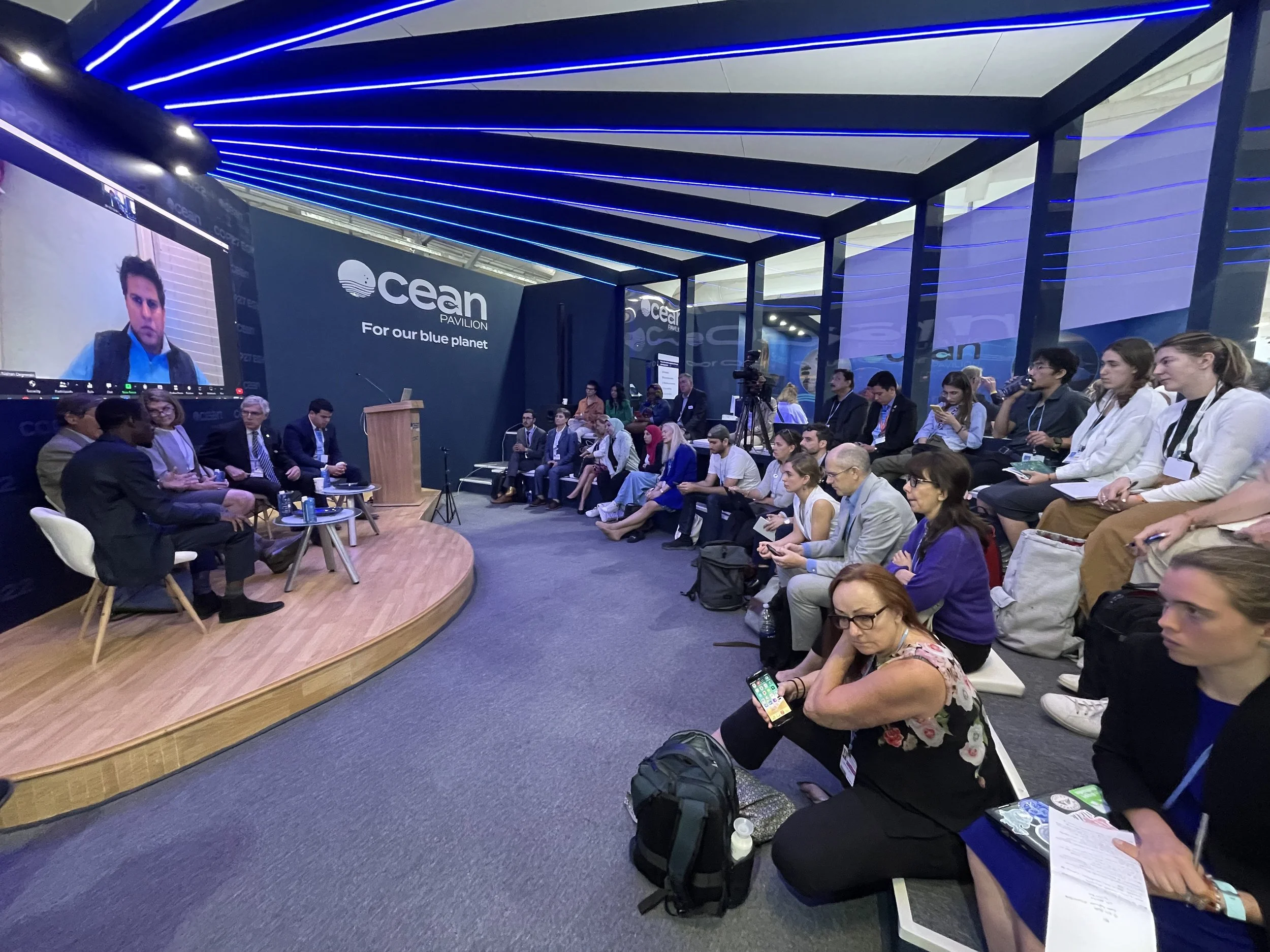 Audience attending a panel discussion at the Ocean Pavilion event with a speaker on stage, large screen, and seating area inside a modern venue.