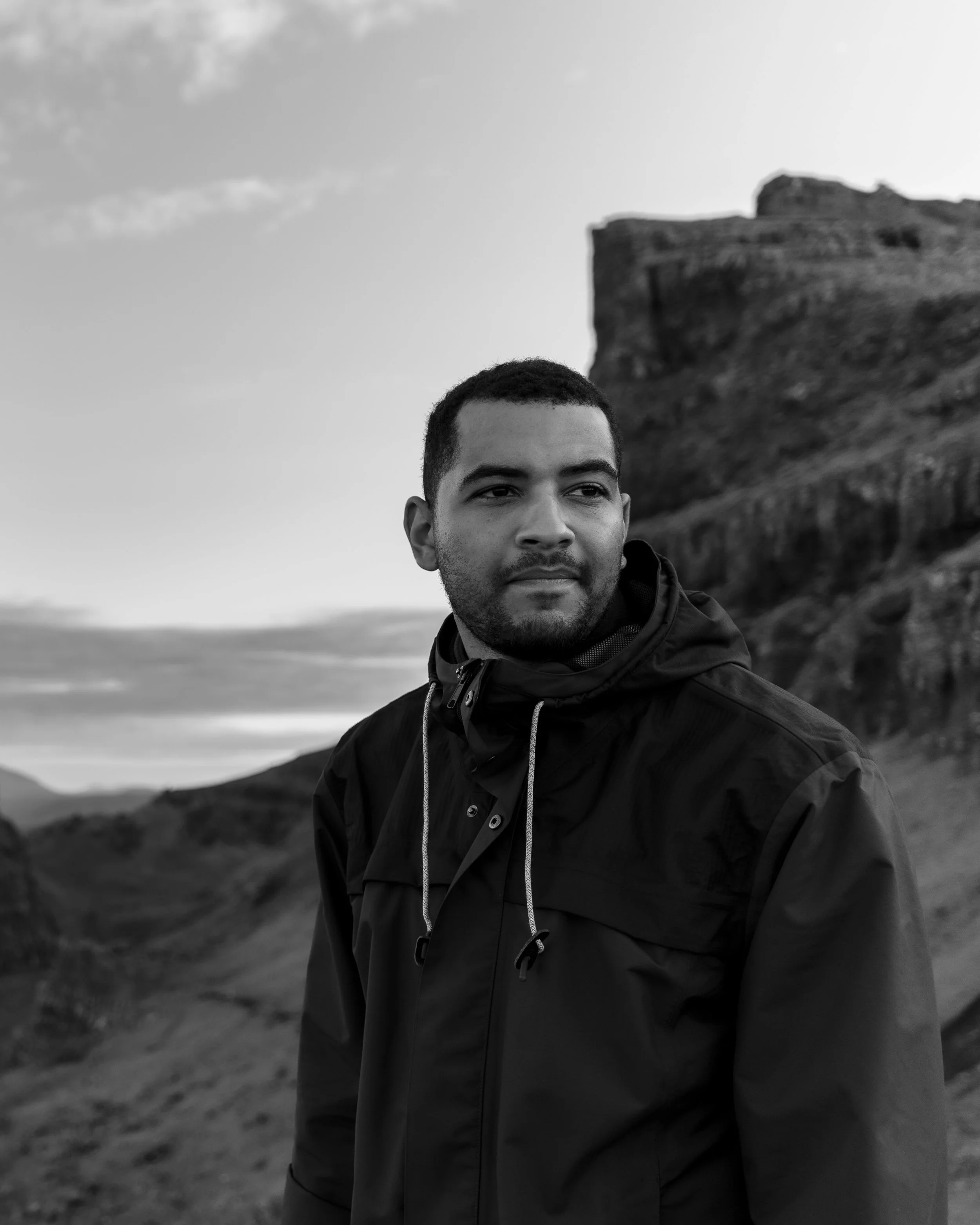 A man in a black jacket standing outdoors with a rocky hill or castle in the background, black and white photo.