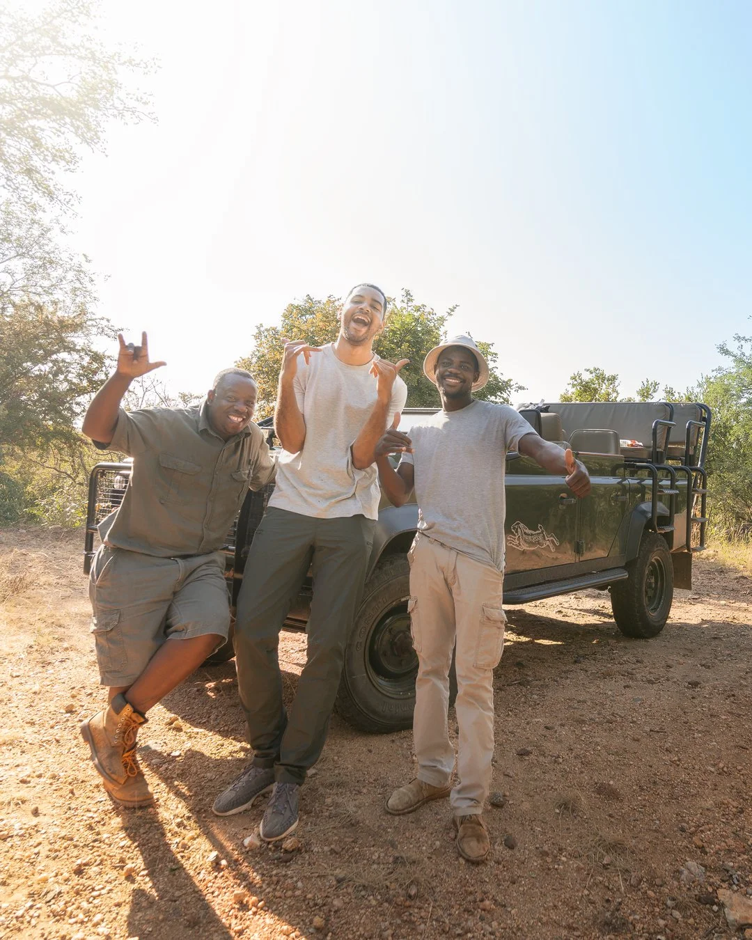 Three men smiling and posing cheerfully outdoors near a black safari vehicle, with trees and a clear sky in the background.
