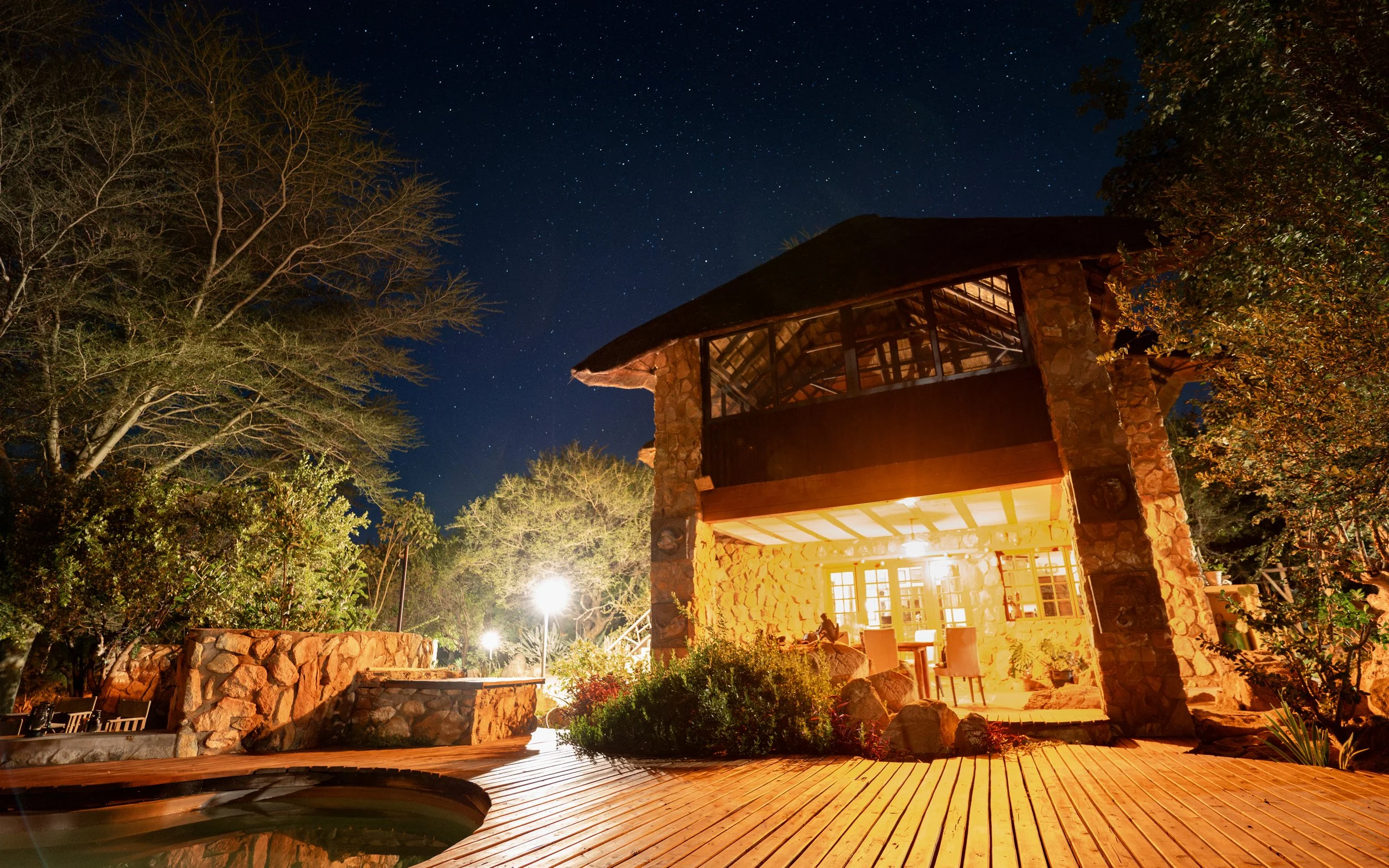 Night view of a two-story house with stone walls and large windows, illuminated from inside, surrounded by trees, with a starry sky overhead and a wooden deck in the foreground.