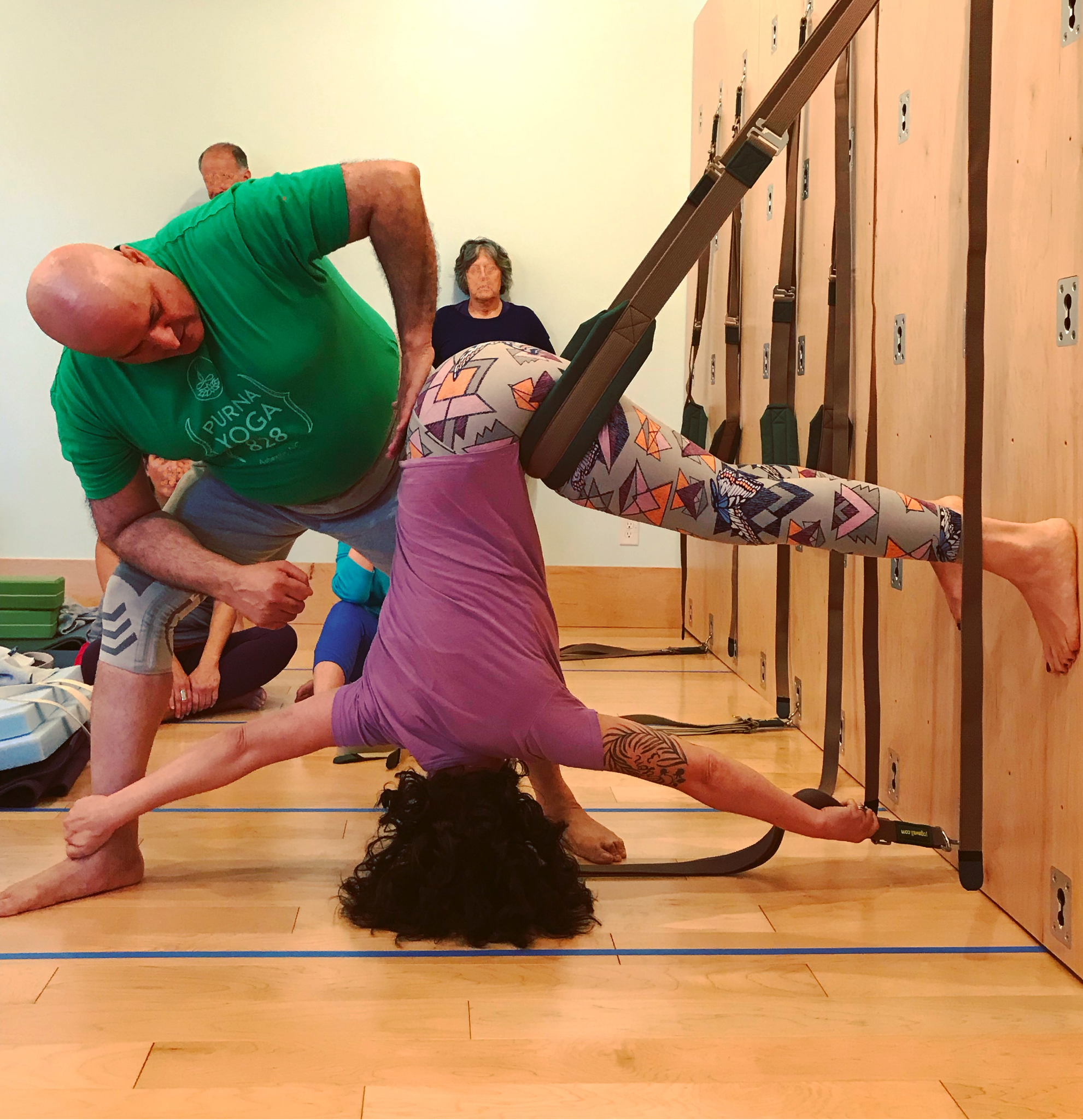 Student in an advanced inverted Great Yoga Wall sling pose with partner support during an immersive yoga wall training