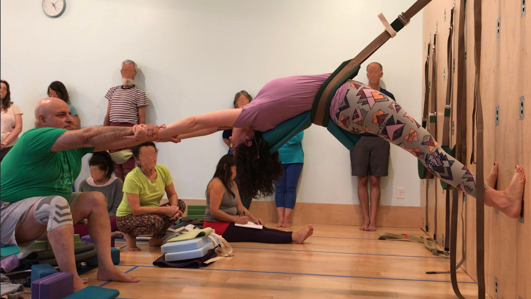 Student in a partner-assisted Great Yoga Wall hip sling forward fold during a large group yoga wall training, observed by fellow trainees
