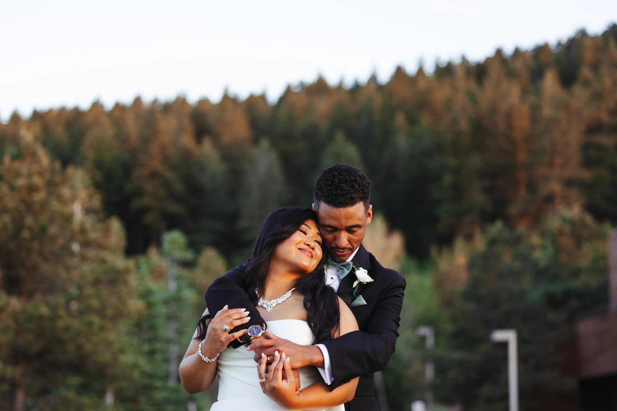 A newlywed couple embracing outdoors with a backdrop of trees, the woman resting her head on the man's shoulder and both smiling contentedly.