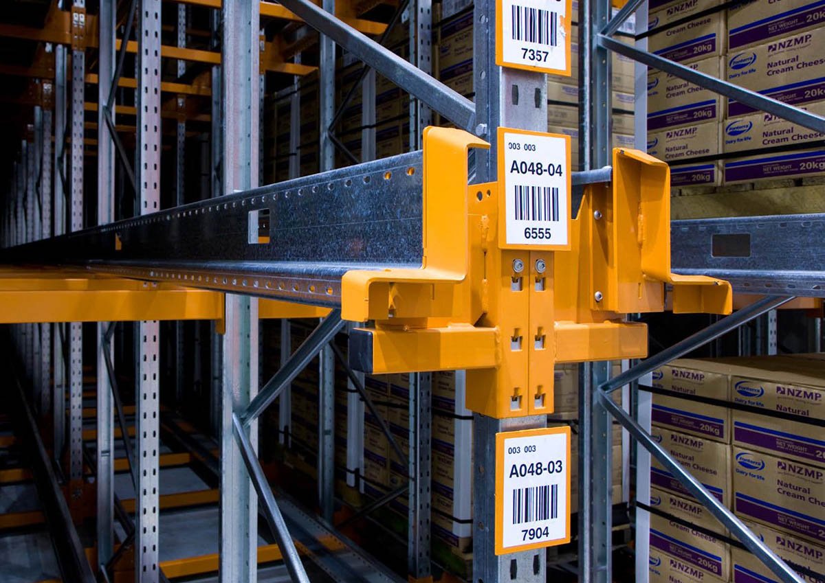Industrial warehouse racking system with orange safety guards and dual barcode location identifiers for inventory tracking
