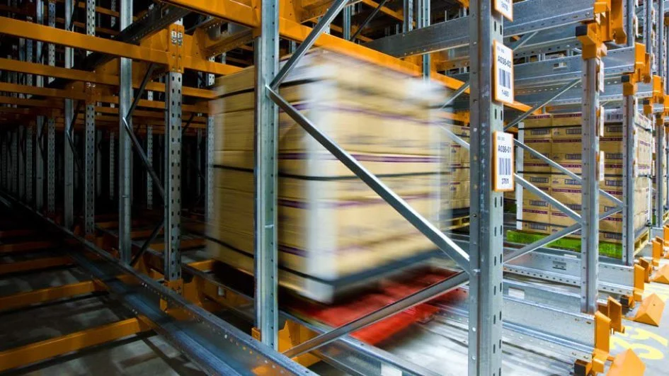 Motion-blurred pallet moving through industrial warehouse racking system with orange safety rails and automated shuttle system