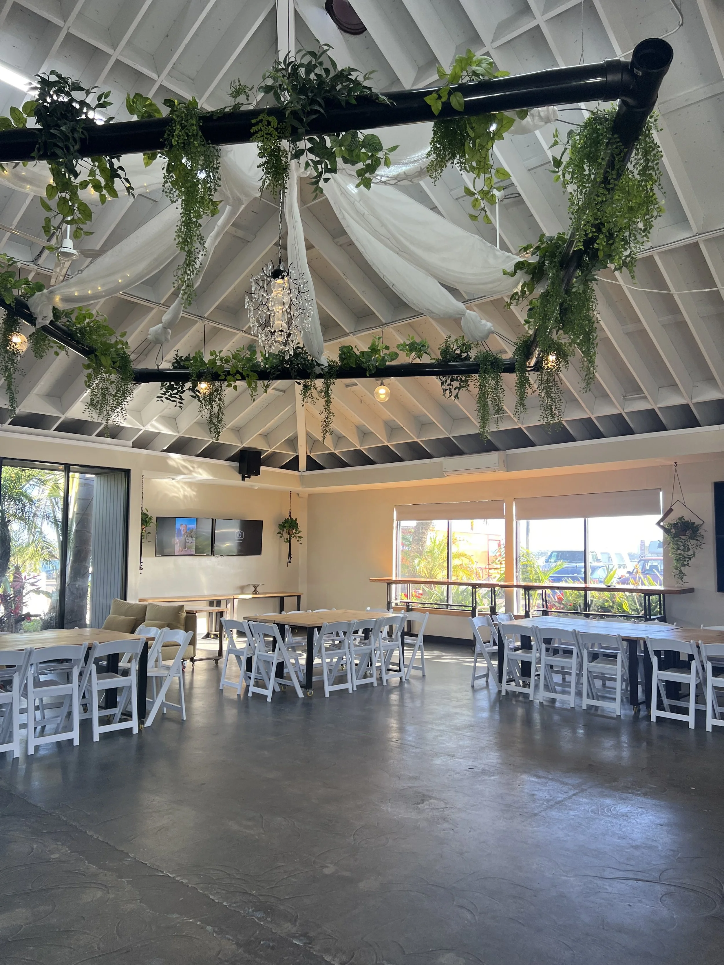Interior of a decorated event space with white chairs and tables, hanging greenery, draped white fabric and a chandelier, with large windows showing outdoor scenery.