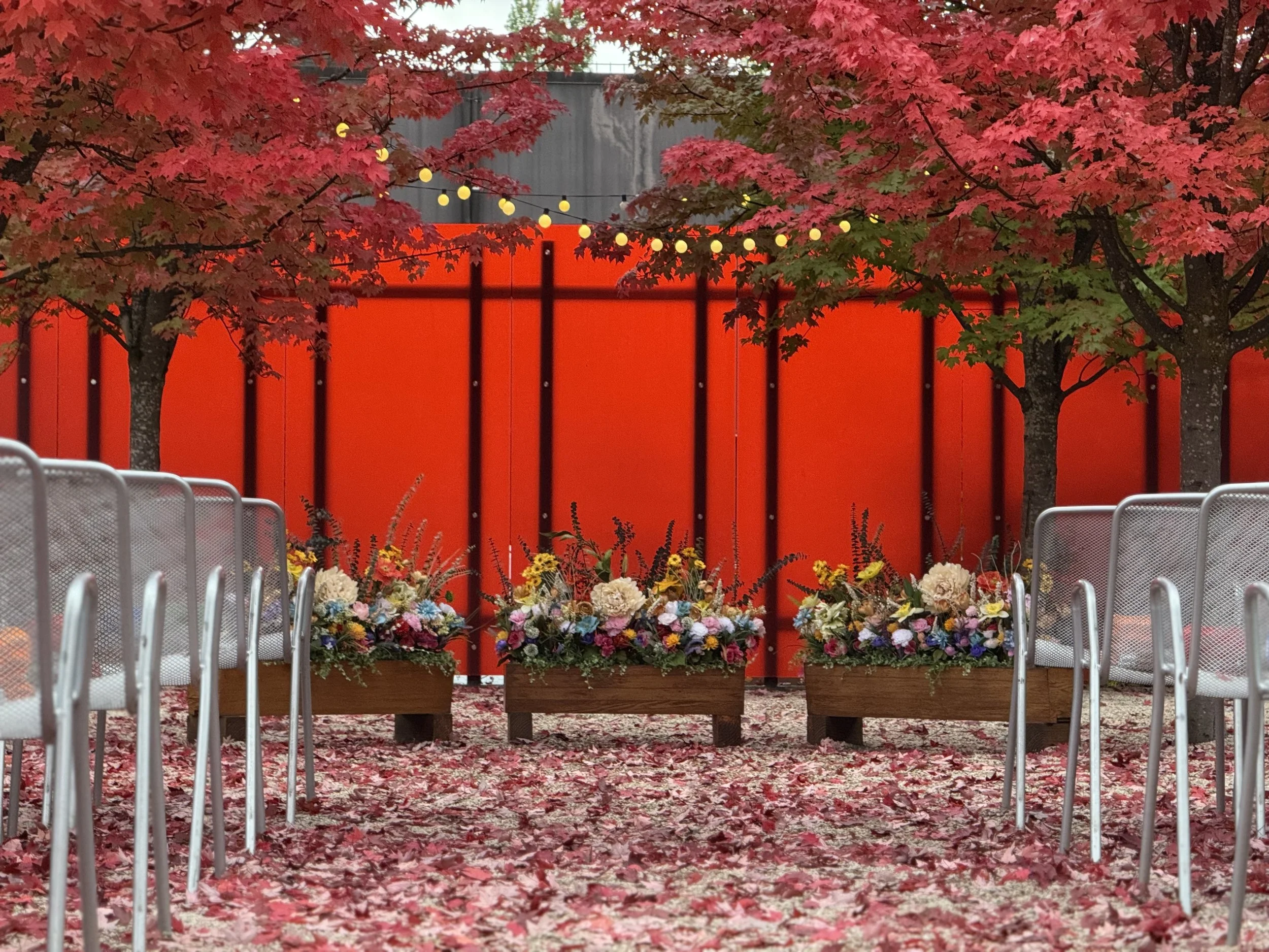 Outdoor ceremony area with rows of metal chairs, three wooden flower boxes filled with colorful flowers, autumn trees with red leaves, a red divider wall, and string lights hanging overhead.