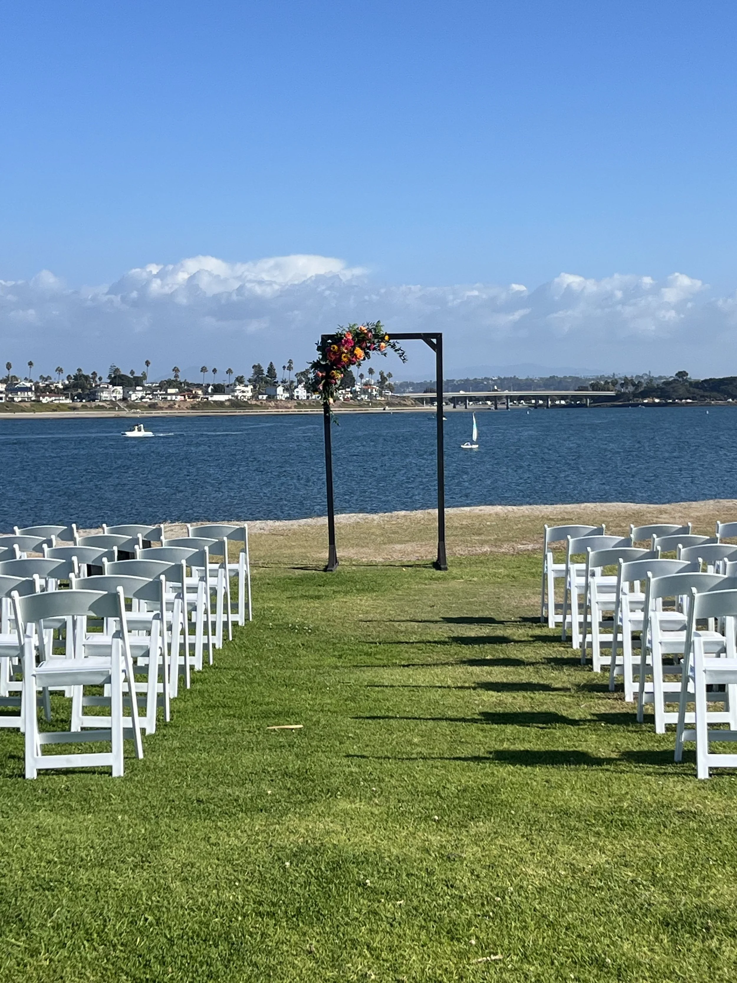 Outdoor wedding setup with white chairs arranged on green grass facing a water with a black metal arch decorated with pink and yellow flowers. Lakeside view with boats and a distant bridge and houses under a partly cloudy sky.