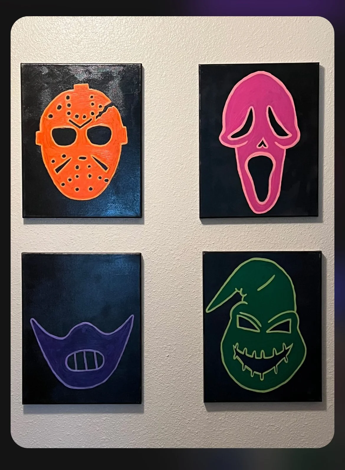 Four colorful Halloween masks displayed on a wall, including a red hockey mask, a pink scream mask, a purple face with sharp teeth, and a green ghostly face with a long nose and jagged mouth.