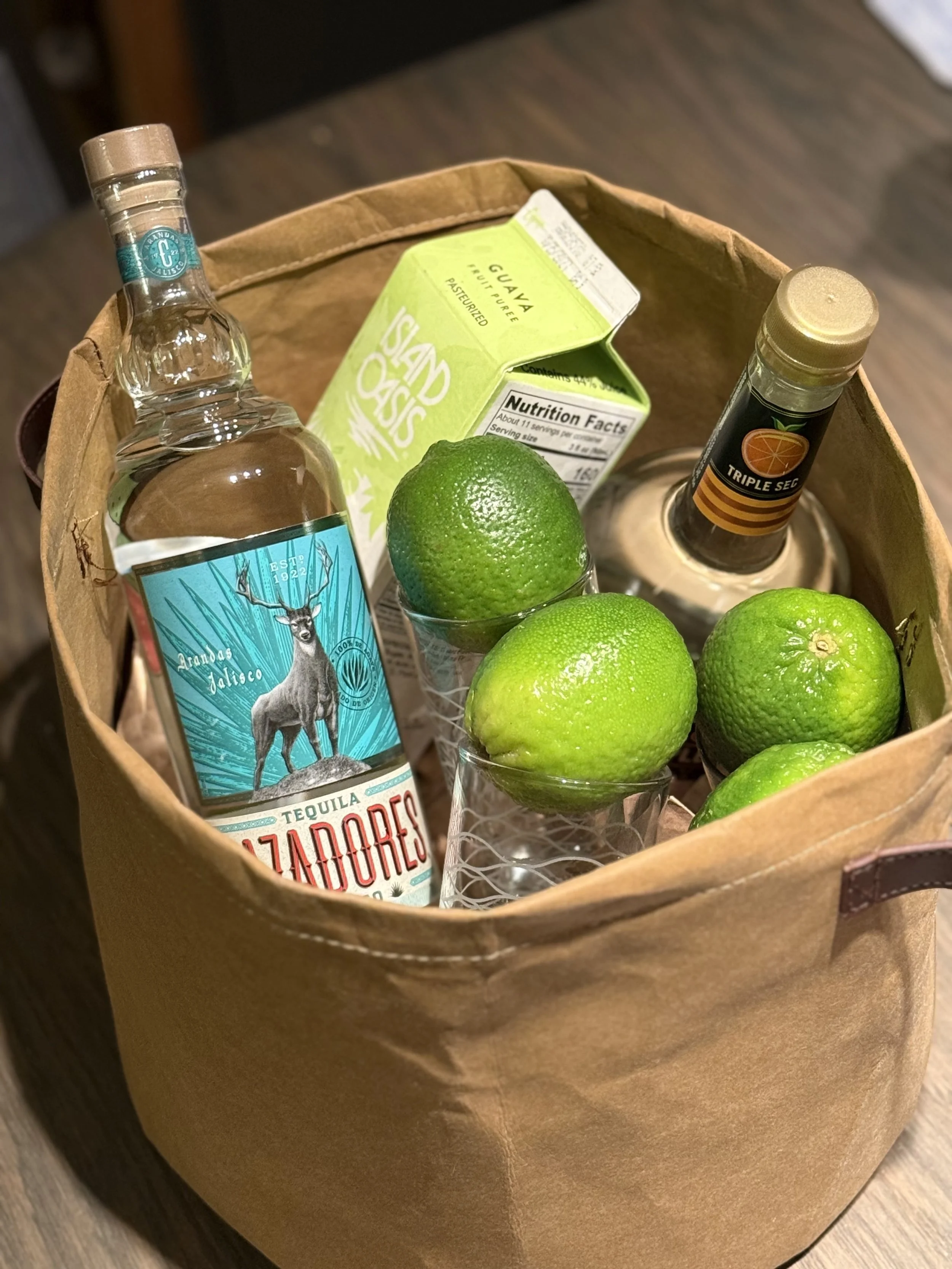 A paper bag containing a bottle of tequila, a lime, a lime-flavored liqueur, a carton of guava juice, and several limes in glasses.