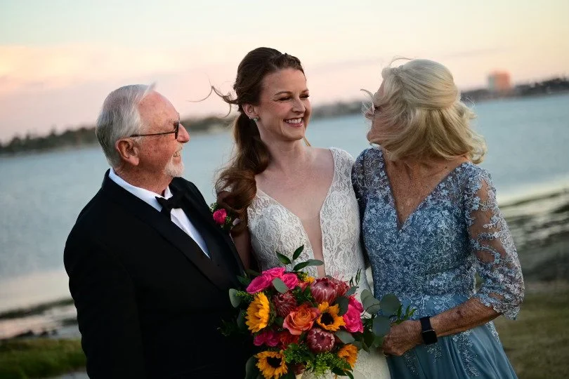 A wedding celebration at the beach during sunset, featuring a bride in a white lace gown holding a colorful bouquet, an older man in a tuxedo, and an older woman in a light blue lace dress sharing a joyful moment.