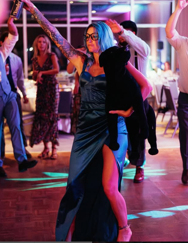 A woman with blue hair, glasses, and tattoos dancing in a dark, colorful party setting, accompanied by a black dog that she holds with her right arm while her left arm is raised.