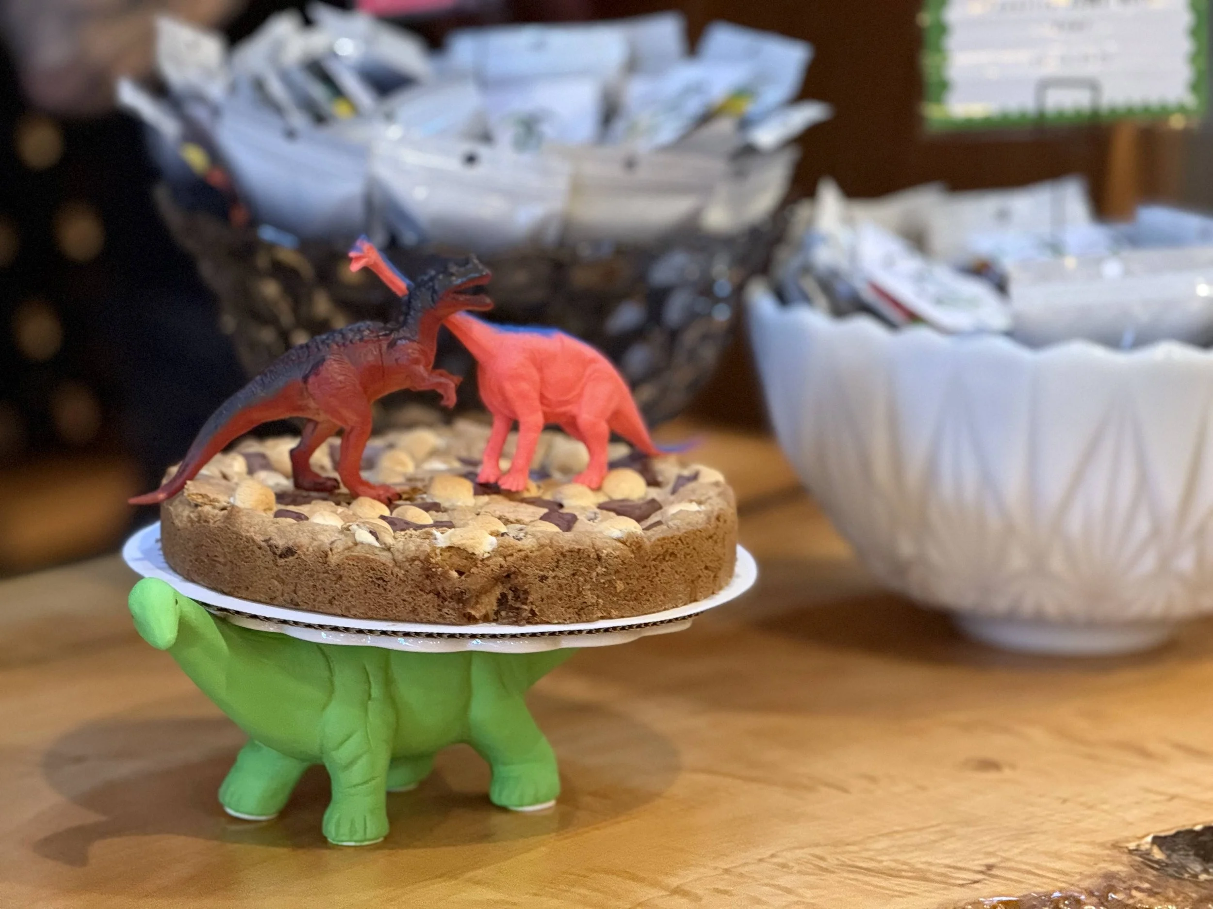 A dessert with two toy dinosaurs on top, placed on a green dinosaur-shaped stand on a wooden table, with bowls and containers of various items in the background.