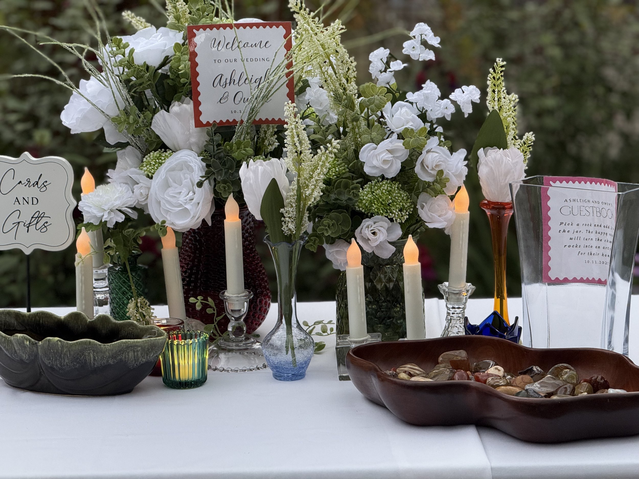 Wedding reception table with white floral arrangements, LED candles, sign cards, and a bowl of polished stones.