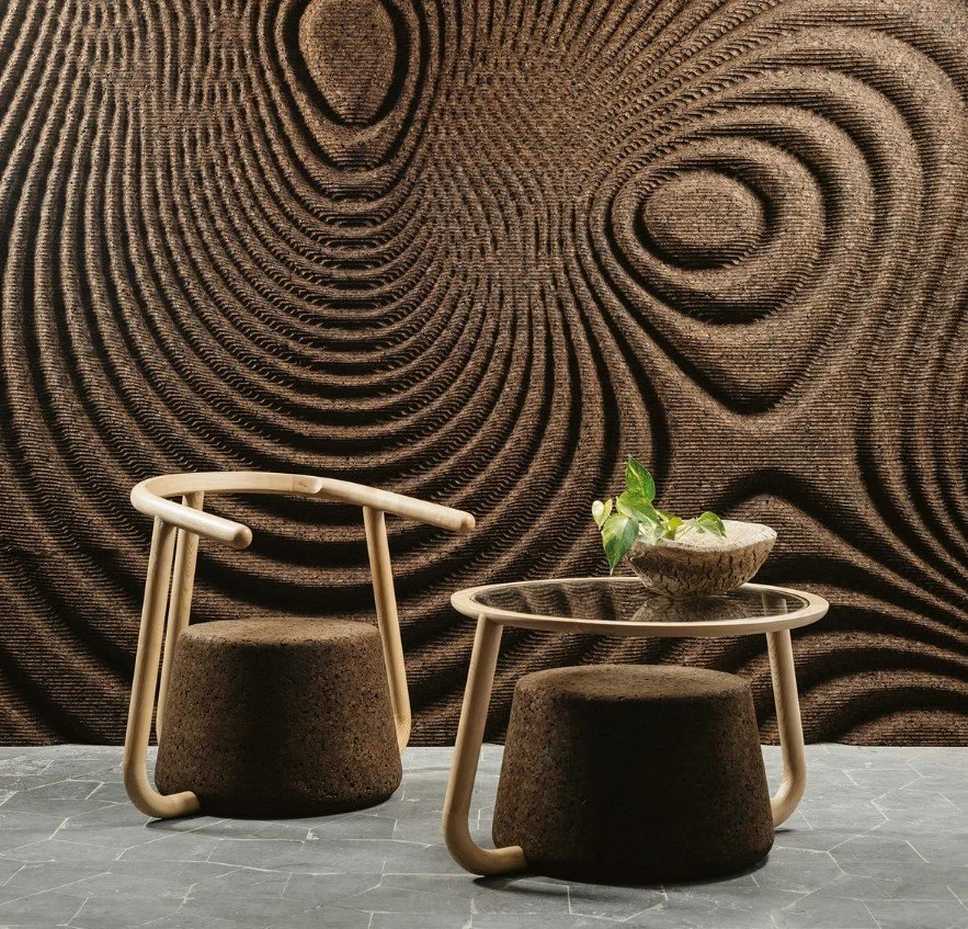 Interior space with a brown textured wall resembling tree rings, a modern wooden chair with a rounded back and dark cushion, a matching stool, and a round table with a bowl containing green leafy plants.
