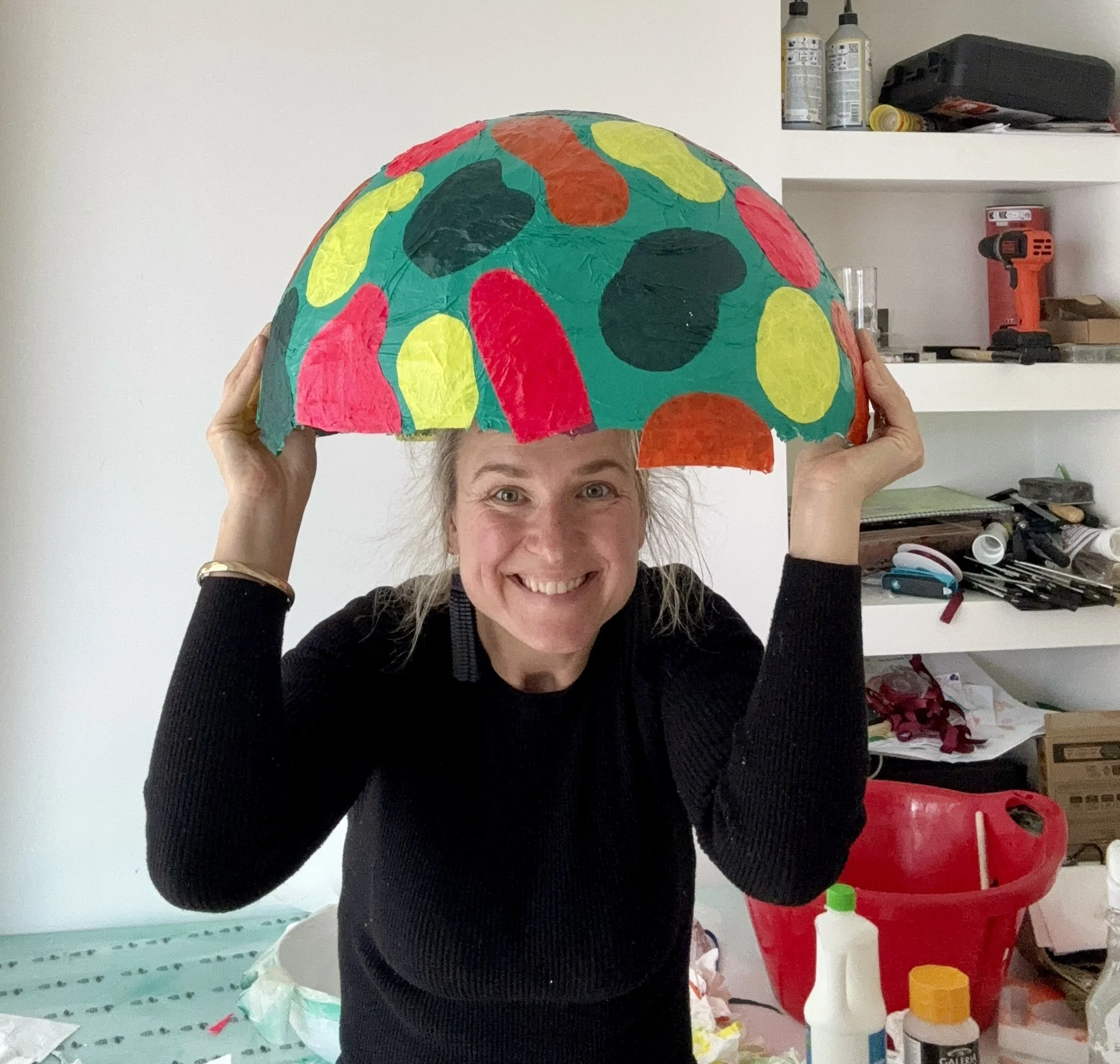 Woman smiling and holding a colorful paper umbrella or hat made of painted paper with red, yellow, black, and orange spots.