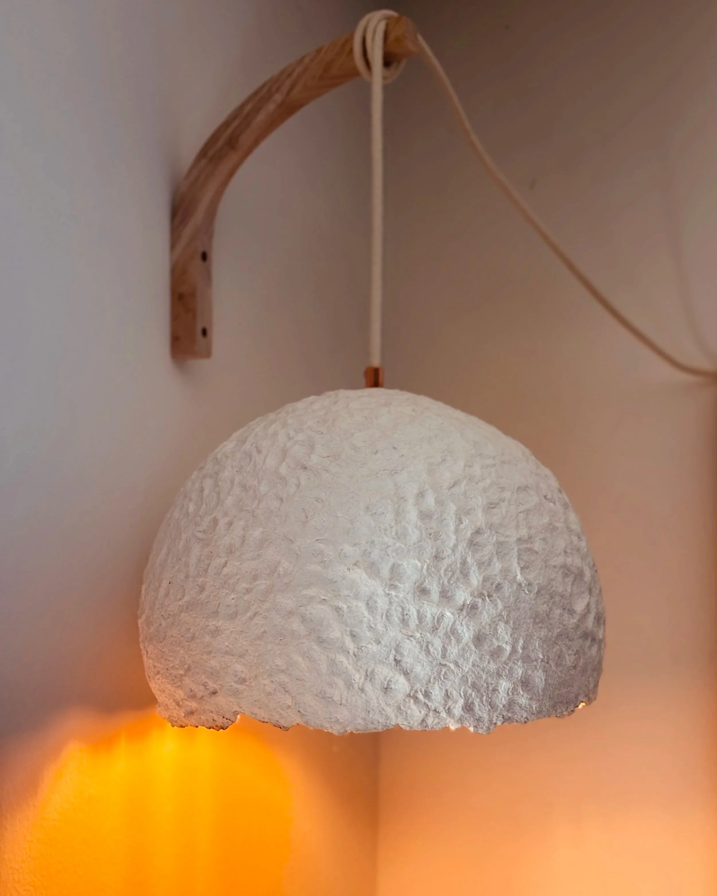 A wall-mounted light fixture with a textured, white, dome-shaped lampshade and a warm glow emanating from beneath, attached to a curved wooden arm.