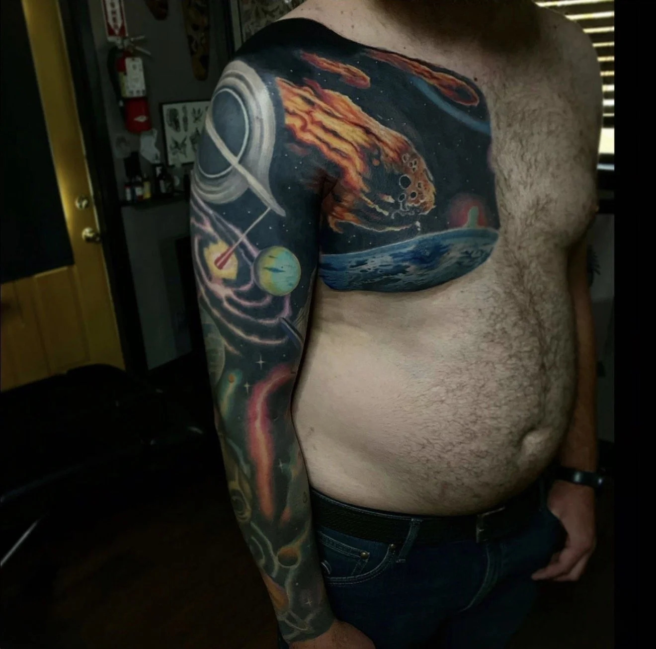Close-up of a man's upper body with detailed space-themed tattoo sleeve covering the left arm and chest, featuring planets, a comet, and cosmic scenery.