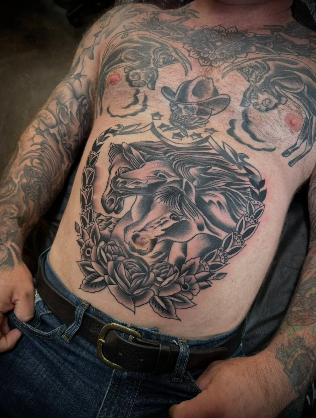 Tattooed man wearing jeans shows tattoos of horses with a floral wreath and a skull wearing a cowboy hat on his chest.