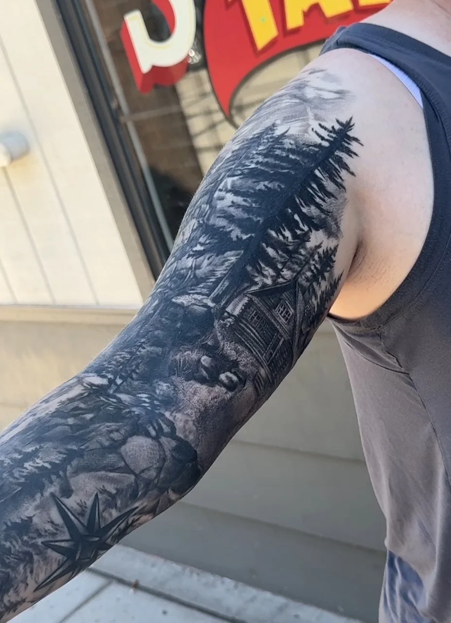 Black and gray tattoo sleeve featuring a mountain cabin, pine trees, and a snow-covered mountain landscape on a person's arm.