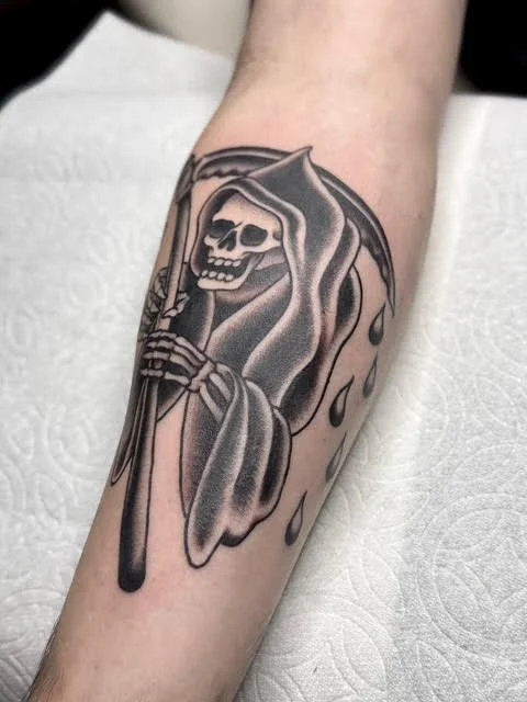 Black and gray trad tattoo of a Grim Reaper skeleton face with a hood, holding a scythe, on a person's forearm.